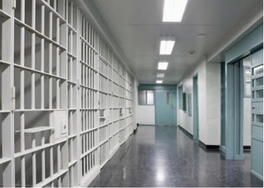 Prison corridor with rows of white jail cells and barred doors along hallway.